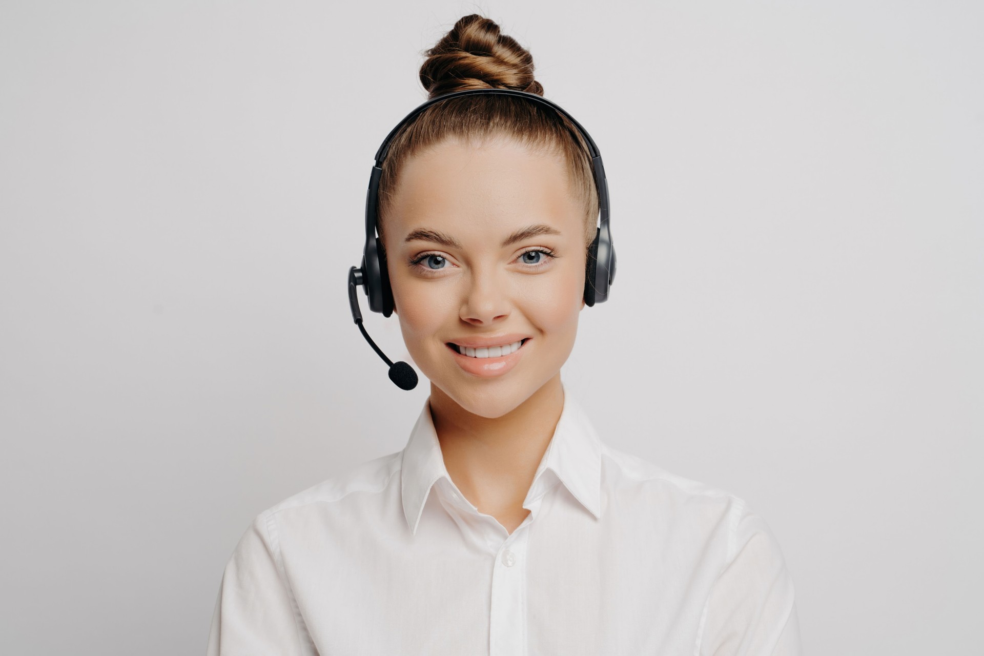 Smiling call center worker in white shirt, hair in bun, black headset, eager to serve and assist customers, stands alone against grey background.
